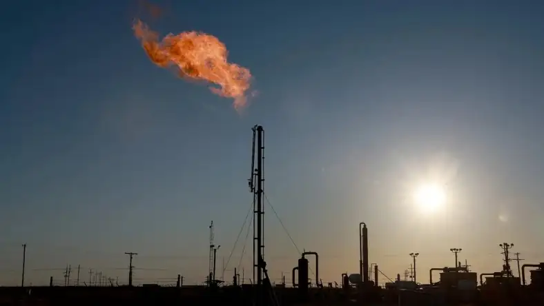 Industrial gas flare burning at dusk with oil refinery infrastructure silhouetted against a pale blue sky